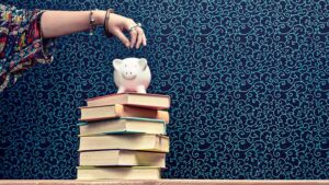 Stack of books with piggy bank on top and person depositing a coin.
