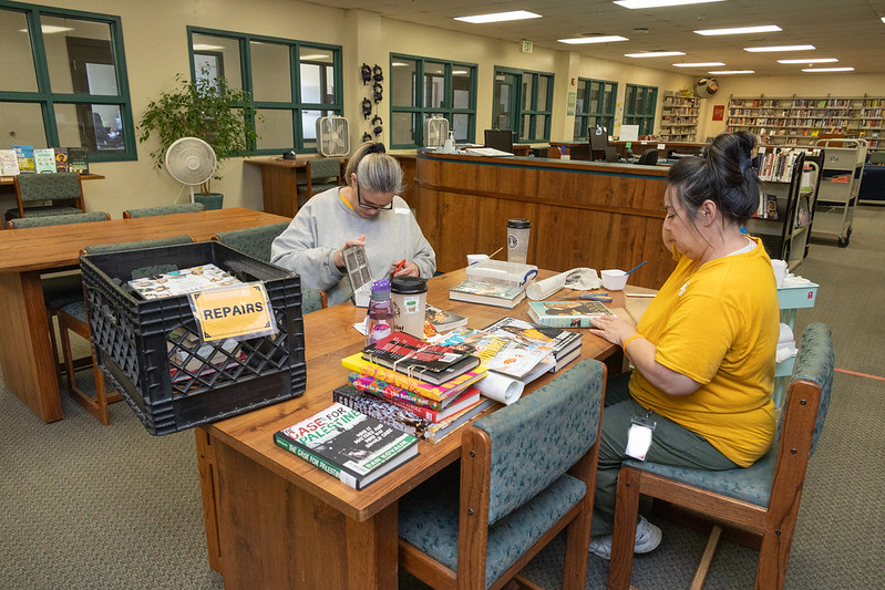 54623072698_8060cb5085_c Two people sitting at a table repairing books in a prison library.