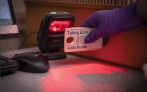 A gloved hand is shown scanning the barcode on a Talking Book cassette.