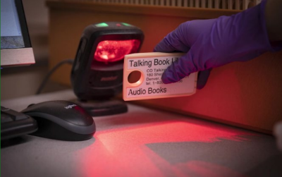 A gloved hand is shown scanning the barcode on a Talking Book cassette.